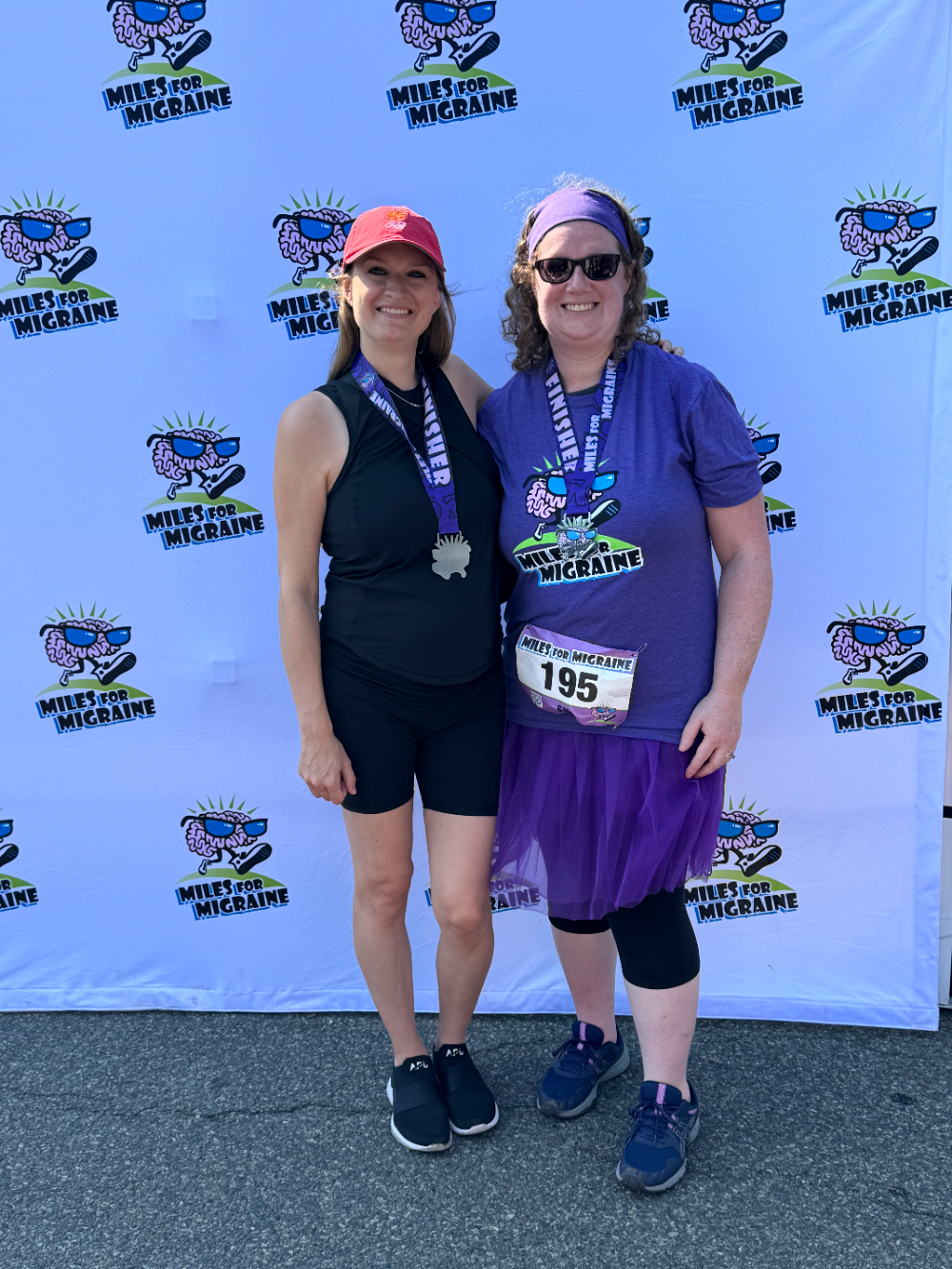 Danielle Newport Fancher with a fellow participant, both wearing finisher medals, in front of a Miles for Migraine backdrop