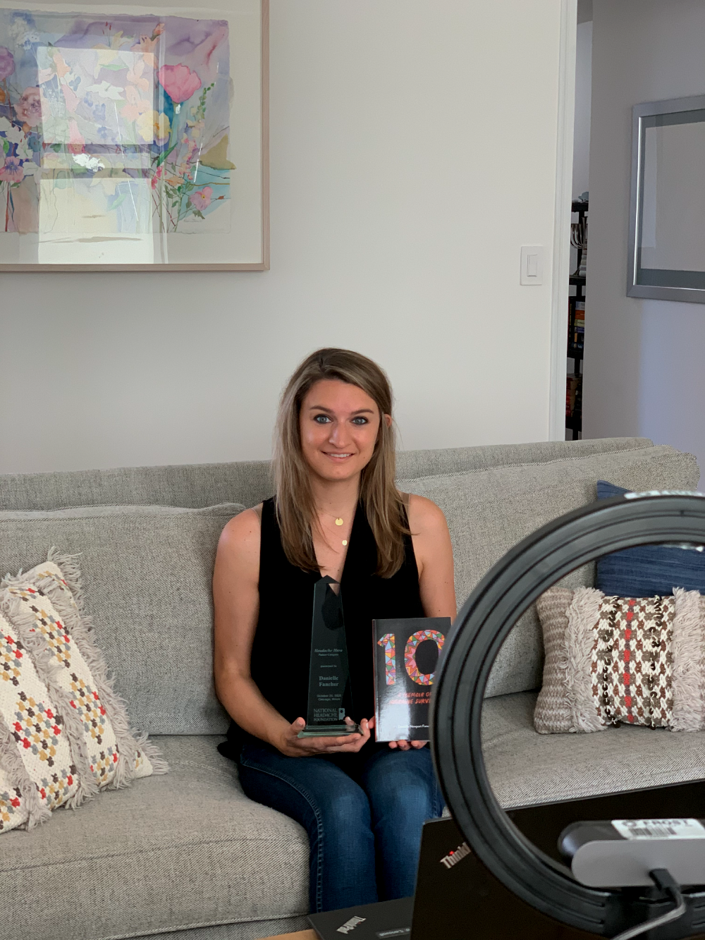 Danielle Newport Fancher seated before a ring light, holding her National Headache Foundation Headache Hero award and a copy of 10
