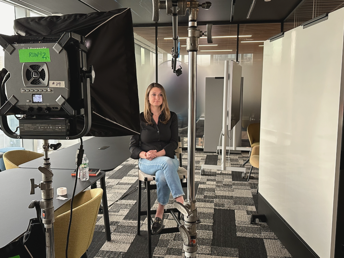 Danielle Newport Fancher seated for a filmed patient-experience interview, softbox lighting and a copy of 10 on the table beside her